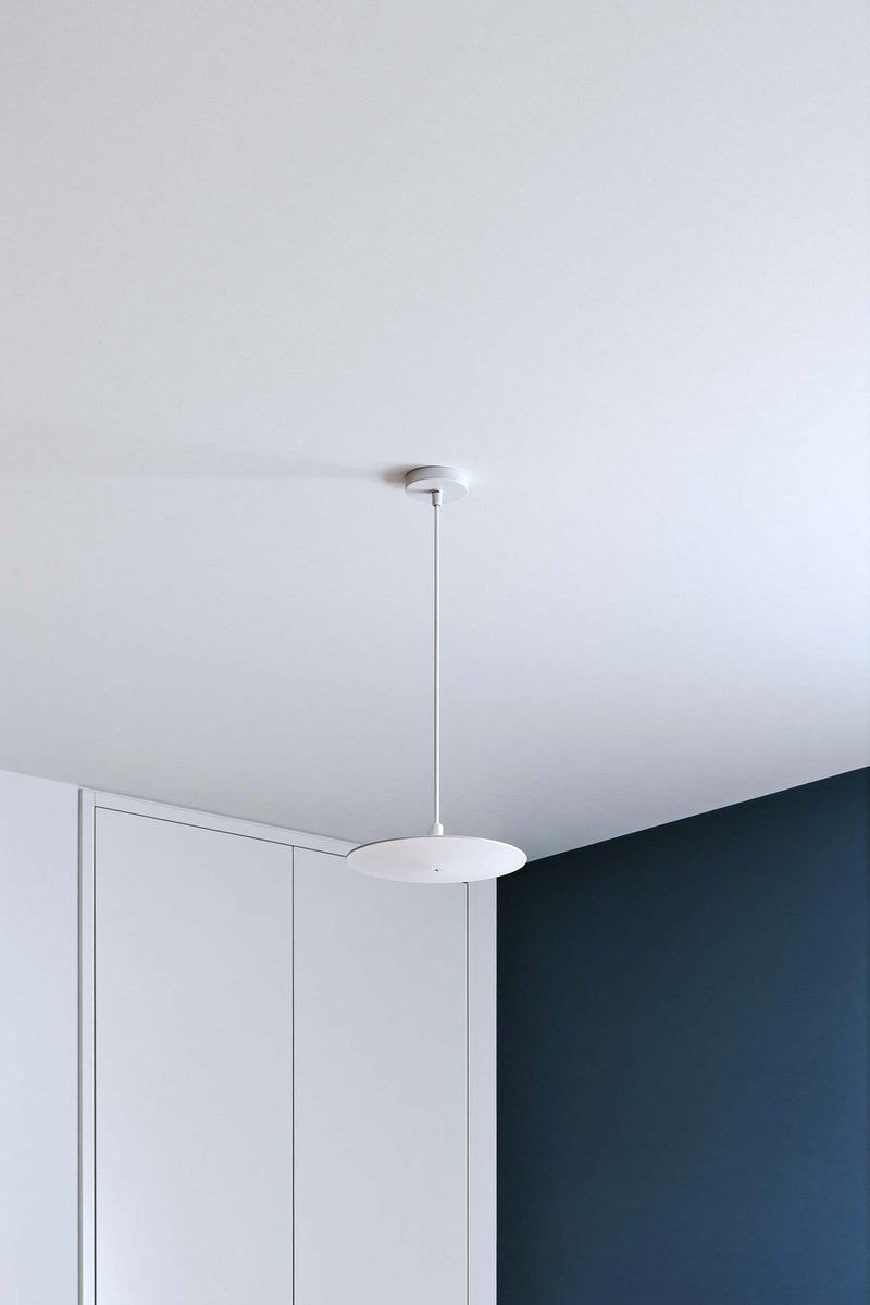 Flat white ceiling seen from below in a furnished bedroom, even and smooth from wall to wall, the tops of tall curtains and a corner of soft greige wall at the edge of the frame
